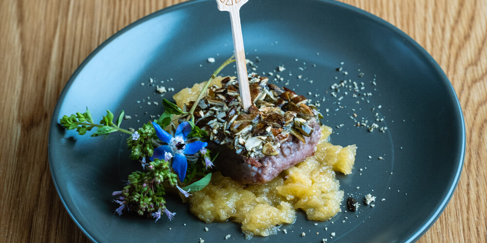 Herb fillet on applesauce, decorated with fresh flowers and herbs on a gray plate, with a small wooden skewer as garnish.